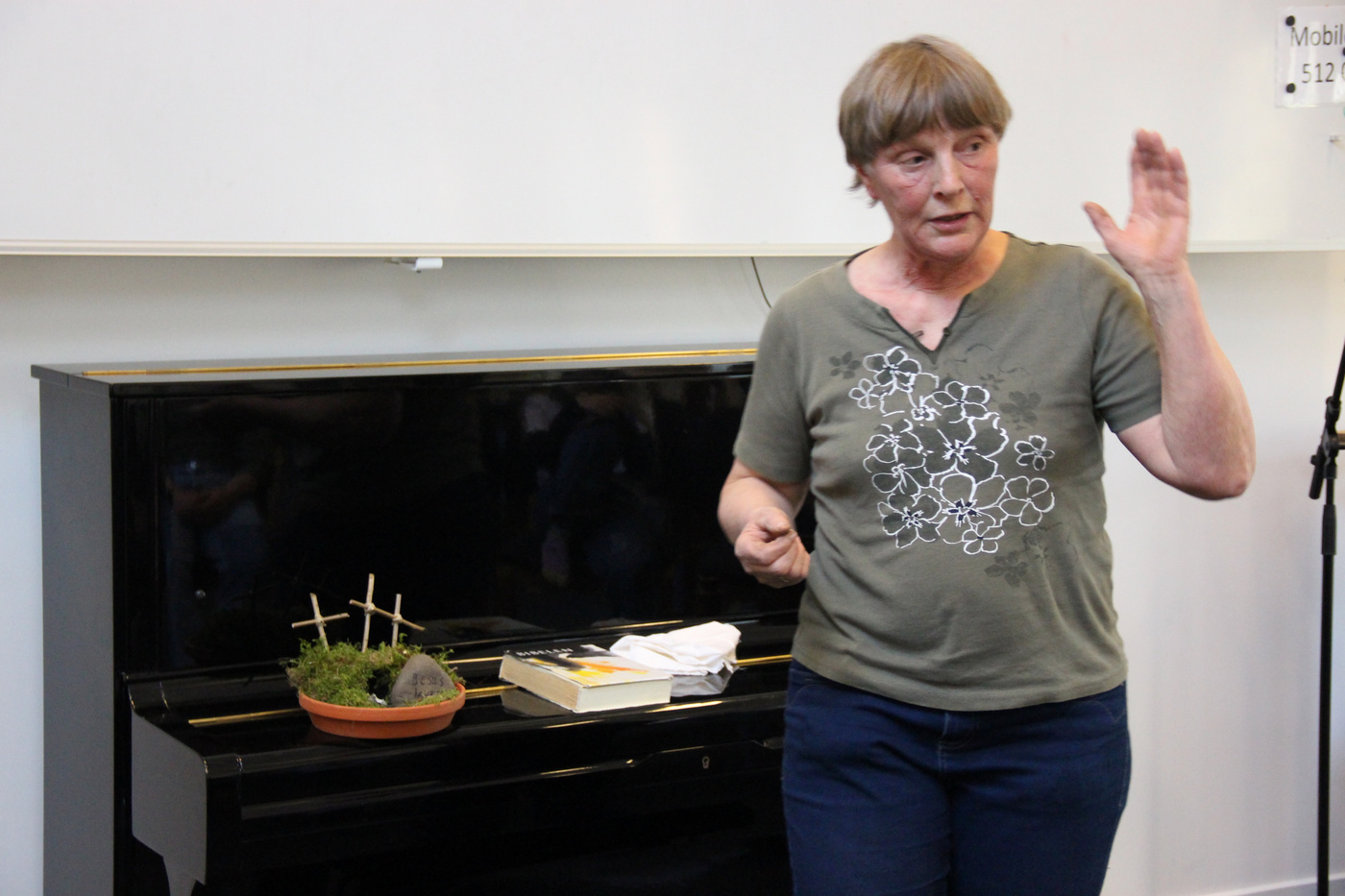 Kvinde gestikulerer, mens hun står ved et klaver med en lille plante.