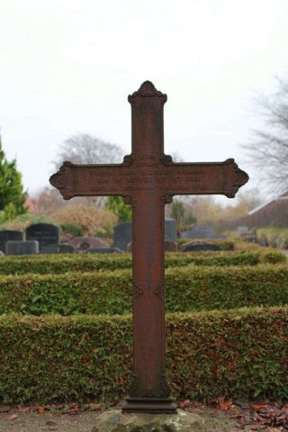 Gammelt, forvitret metalkors på en kirkegård med grønne hække.