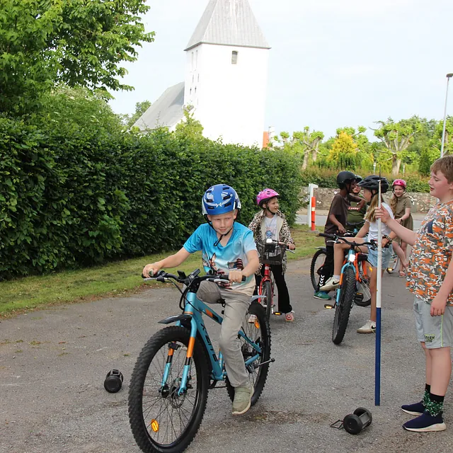 Børn på cykler, nogle med hjelme, nær en kirke om sommeren.