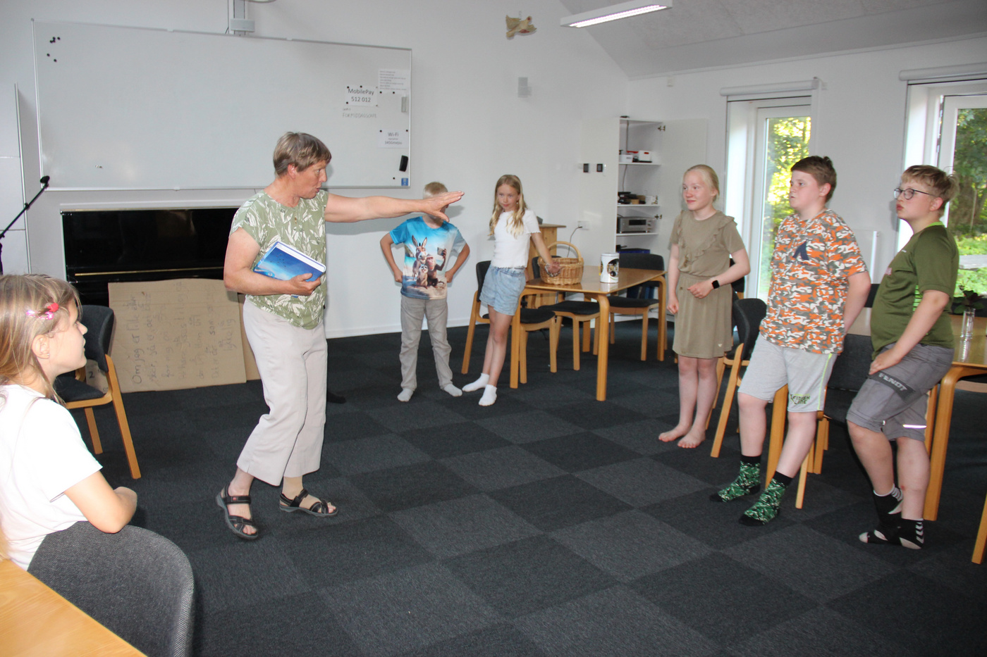 Kvinde leder en gruppe børn i en aktivitet i et lyst rum.