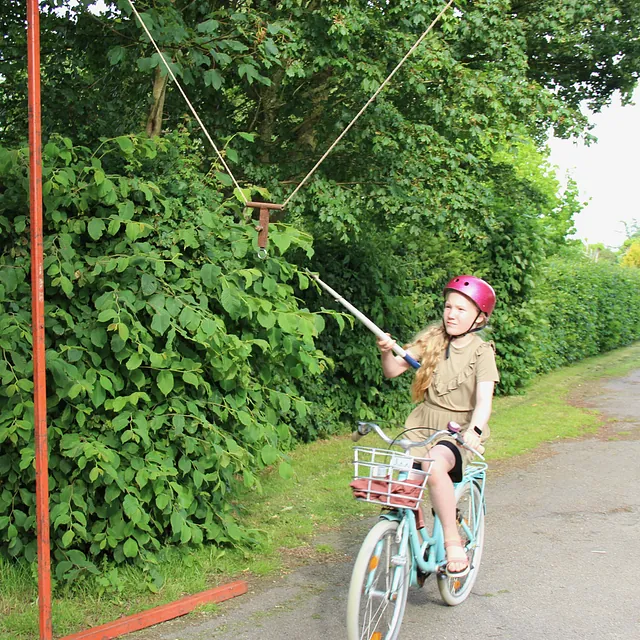 Pige på cykel, med hjelm, rækker ud efter ophængt genstand med pind.