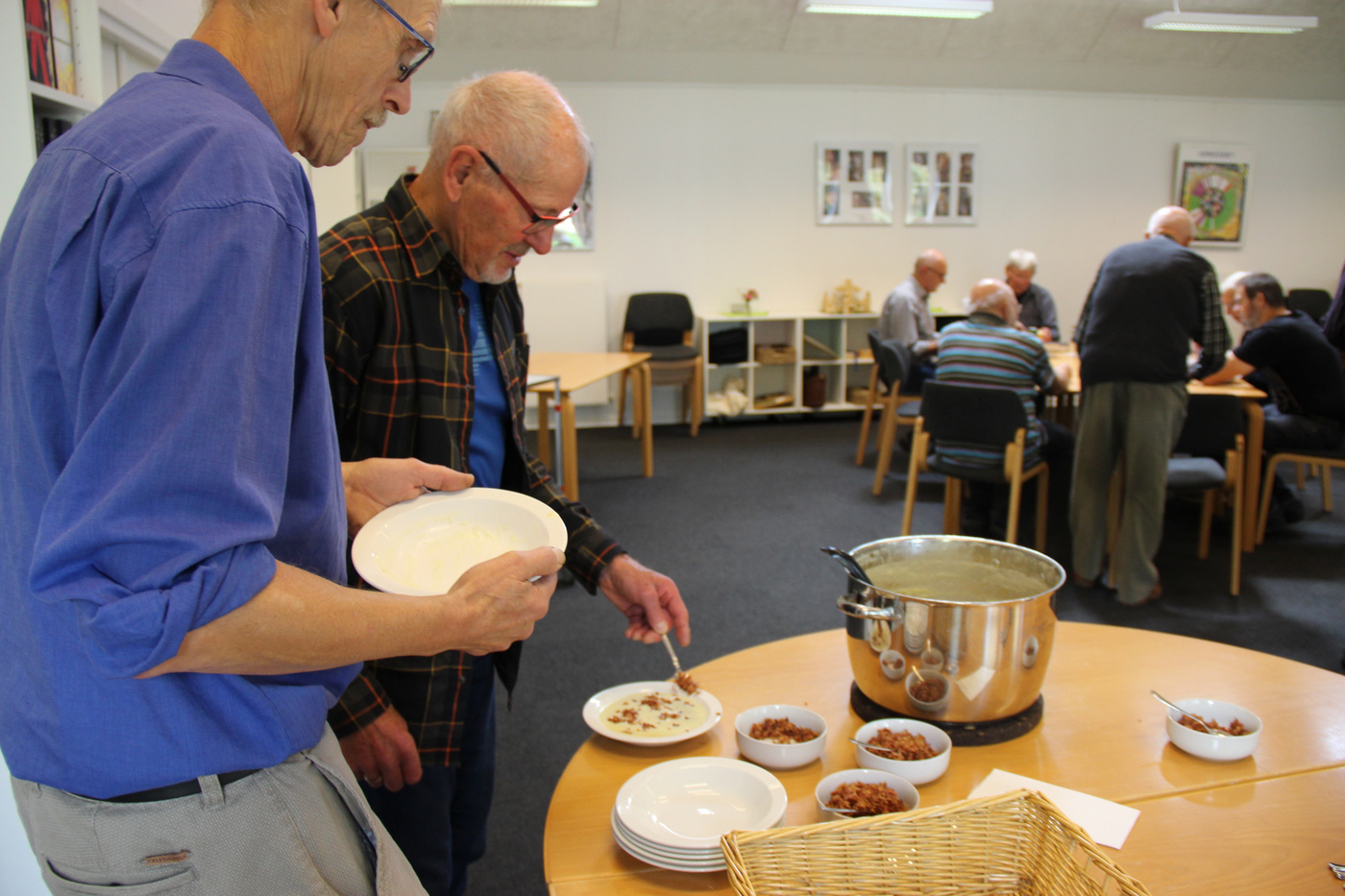 To mænd serverer mad fra en stor gryde til skåle på et bord.