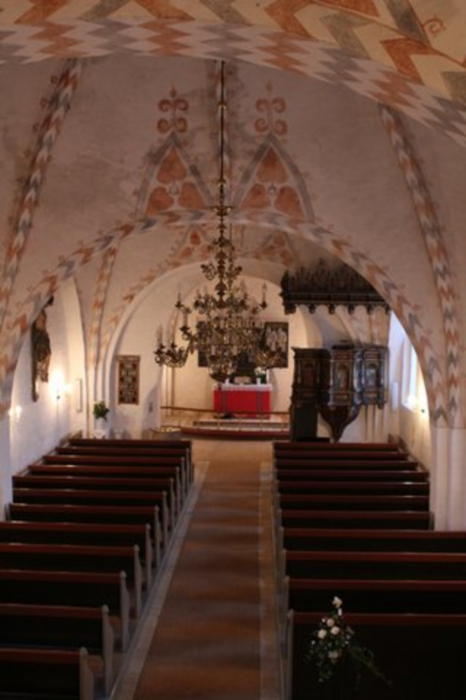Kirkeinteriør med mønstret loft, kirkebænke, alter og lysekroner.