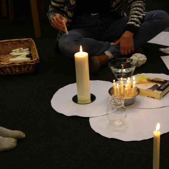 Tændte lys, bibel og kurv med en persons ben synlige på gulvet.