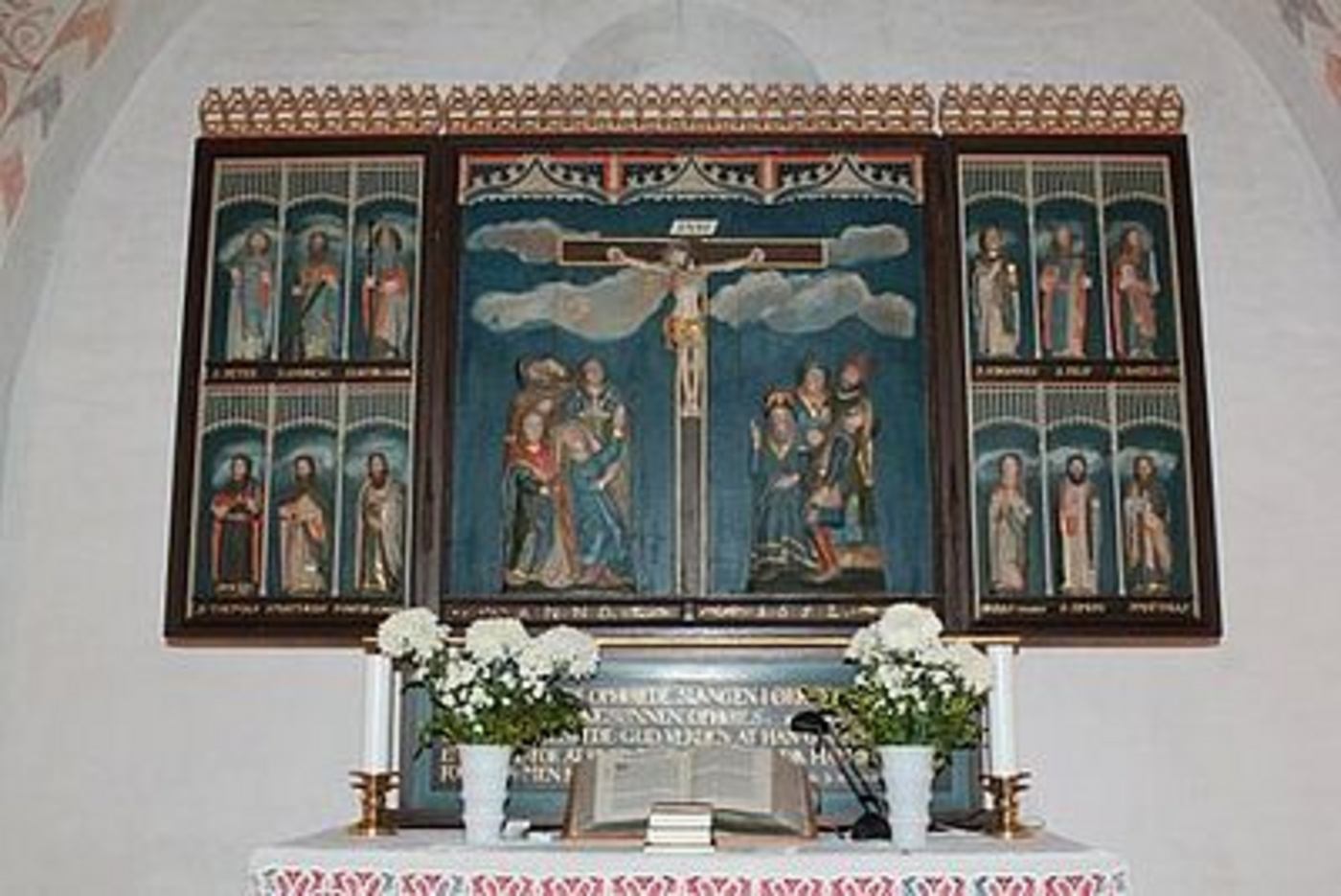 Detaljeret altertavle i træ med korsfæstelsesscene, blomster og åben bog.