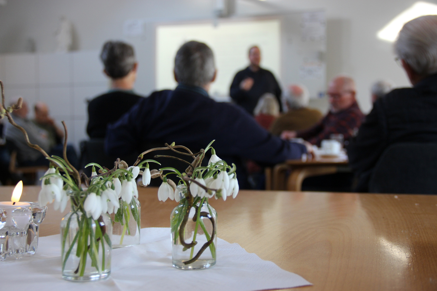 Vintergækker og lys på bord, sløret publikum vender mod en taler.
