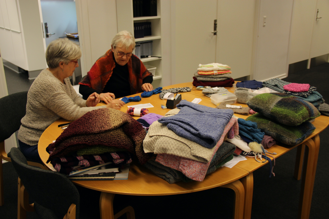 To kvinder sorterer strikkede beklædningsgenstande på et bord i et lyst rum.
