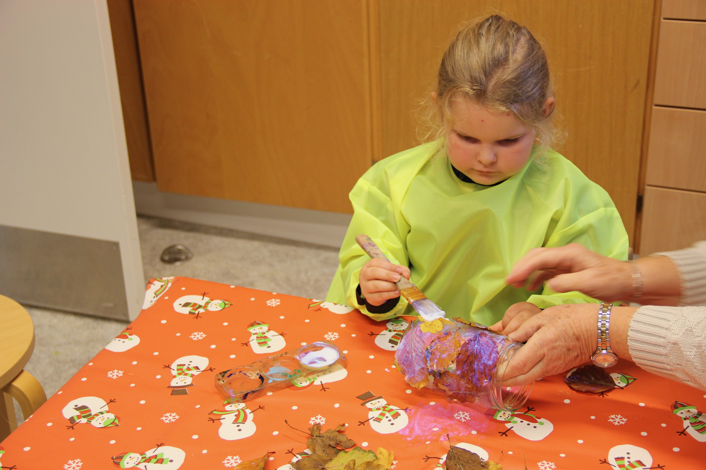 Barn i kittel maler håndværk, voksen assisterer ved et bord.