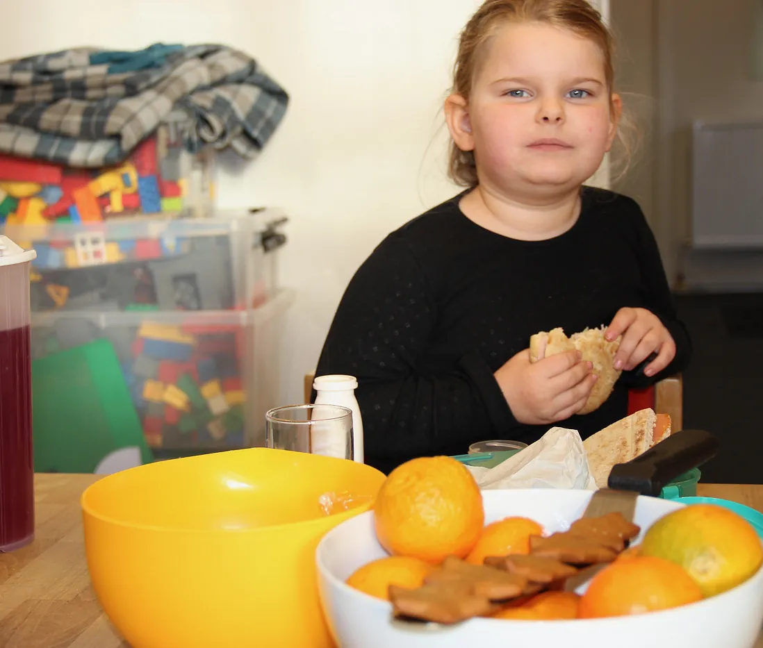 Barn spiser sandwich ved bord med frugt og legetøj.