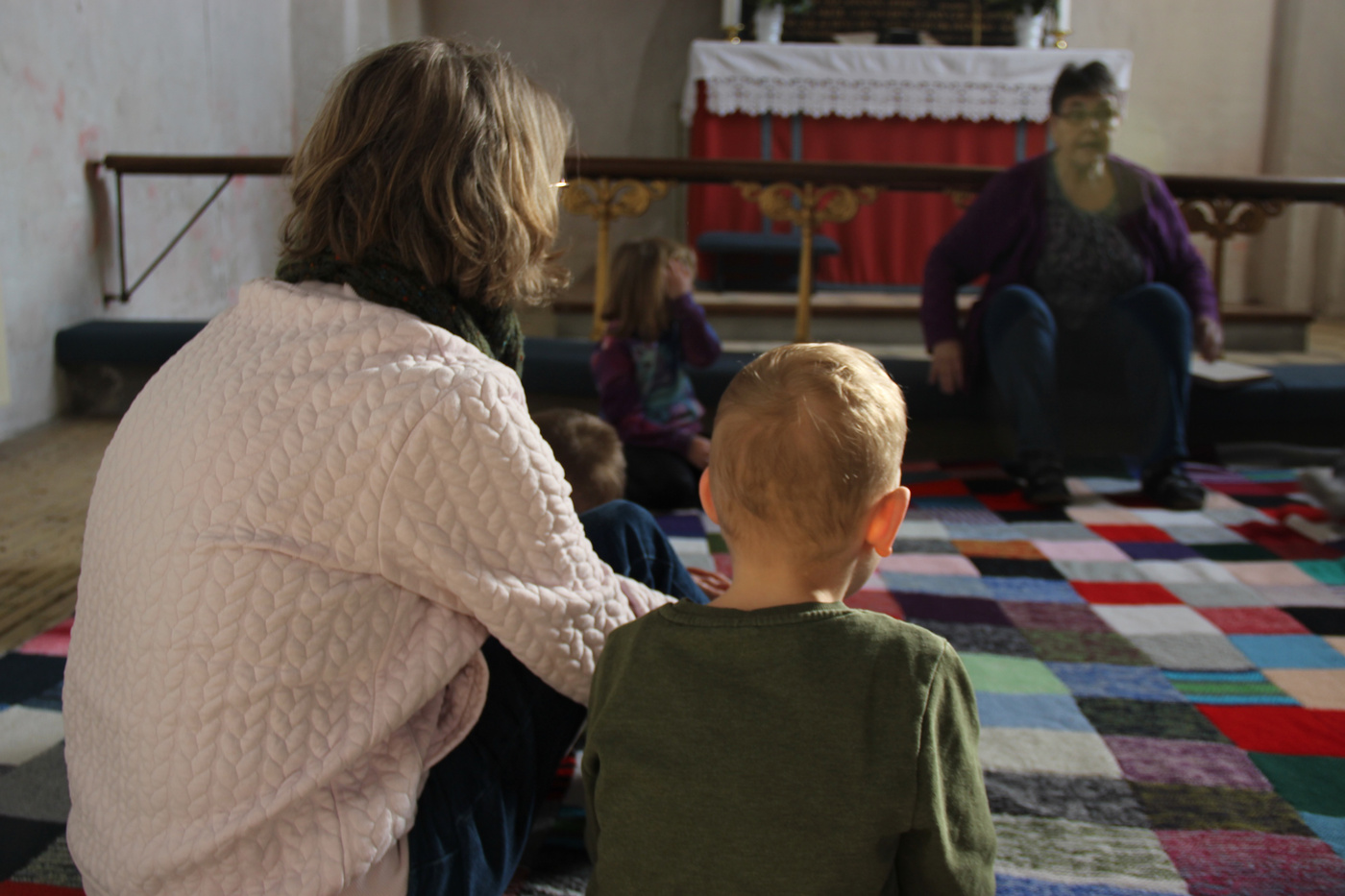 Kvinde og barn sidder på et farverigt tæppe i kirken og lytter.