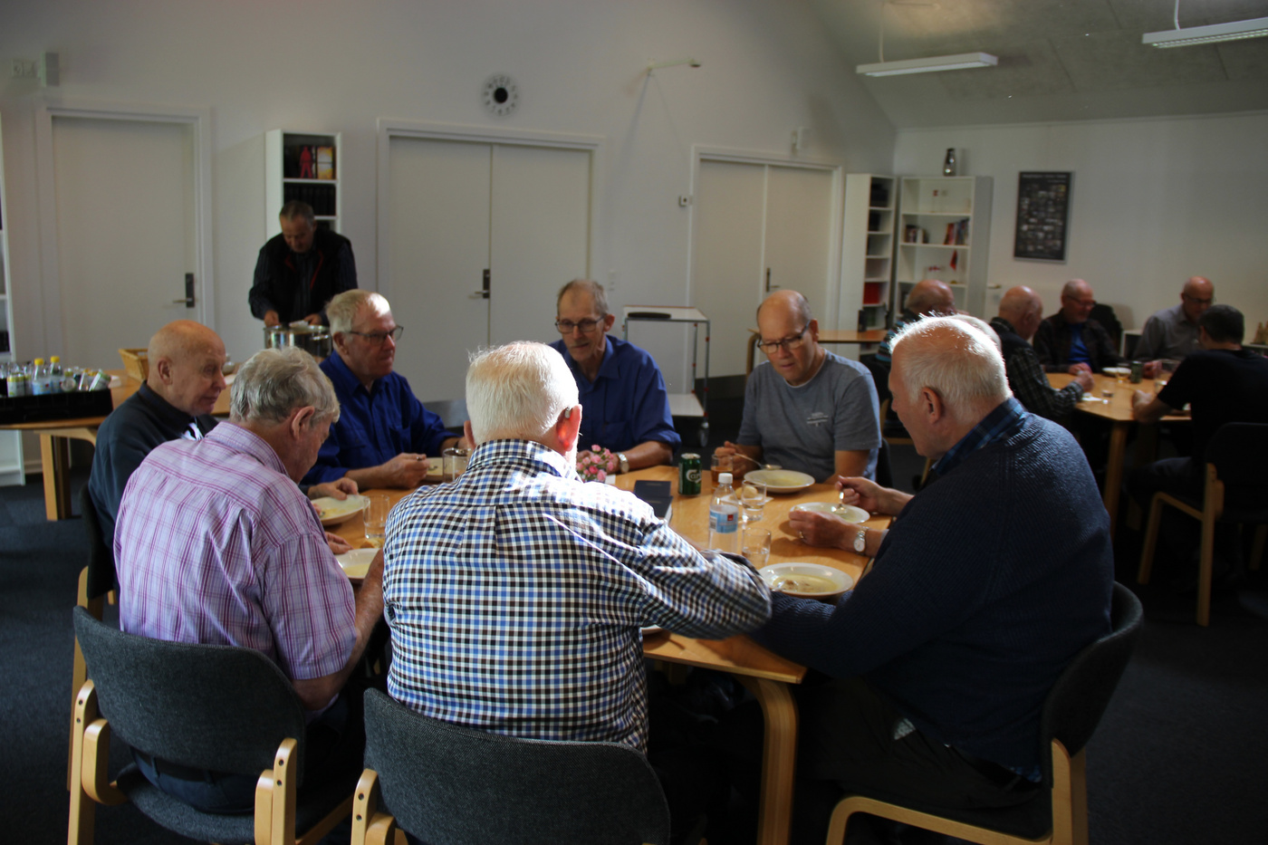  Flere ældre mænd spiser og snakker ved borde i et lyst rum.