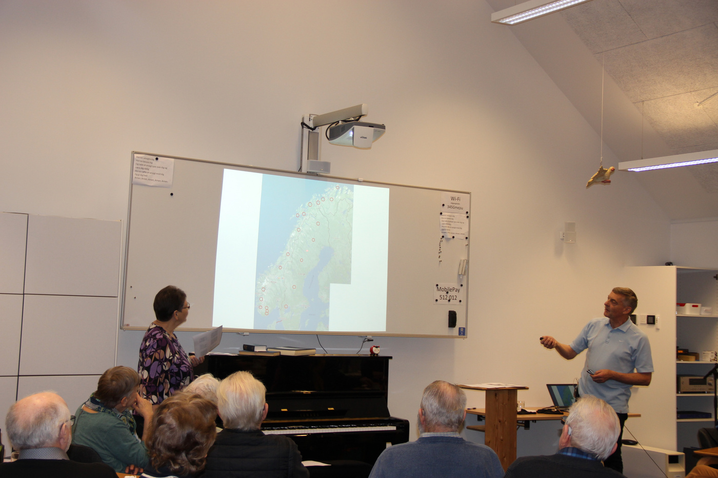 To præsentanter diskuterer et kort projiceret på en skærm for et publikum.