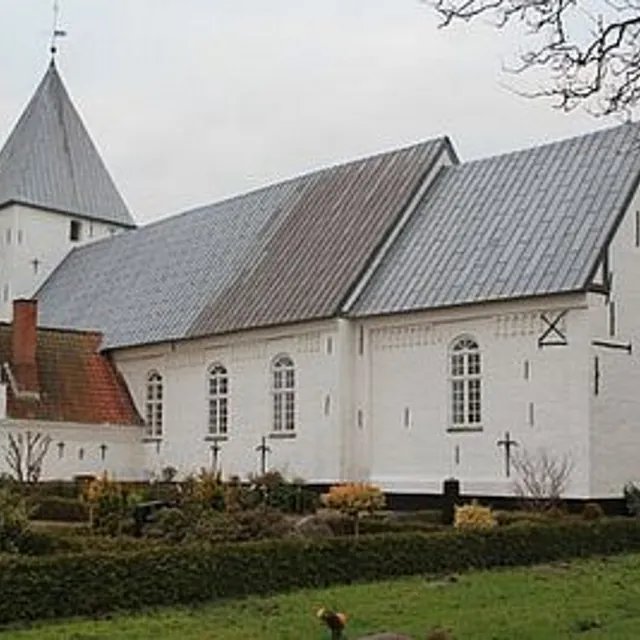 Hvid kirke med gråt tag og spir, omgivet af grønne buske.