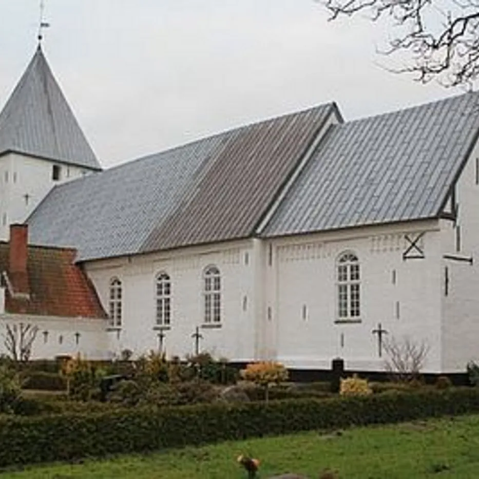 Hvid kirke med gråt tag og spir, omgivet af grønne buske.