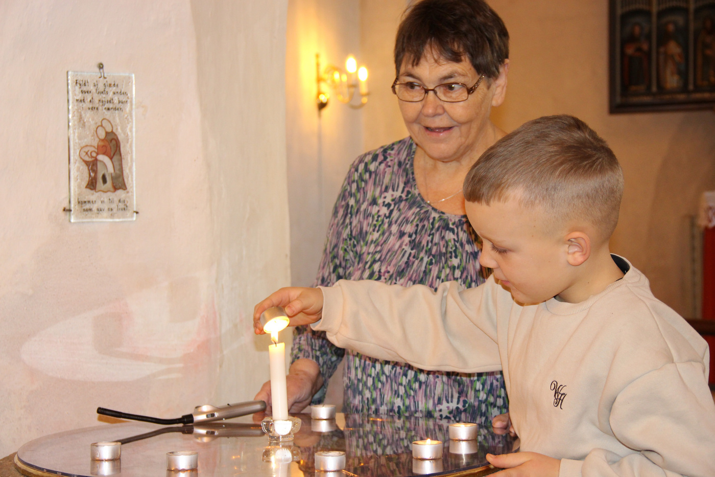 Dreng tænder et lys ved en stenfont, observeret af en voksen.