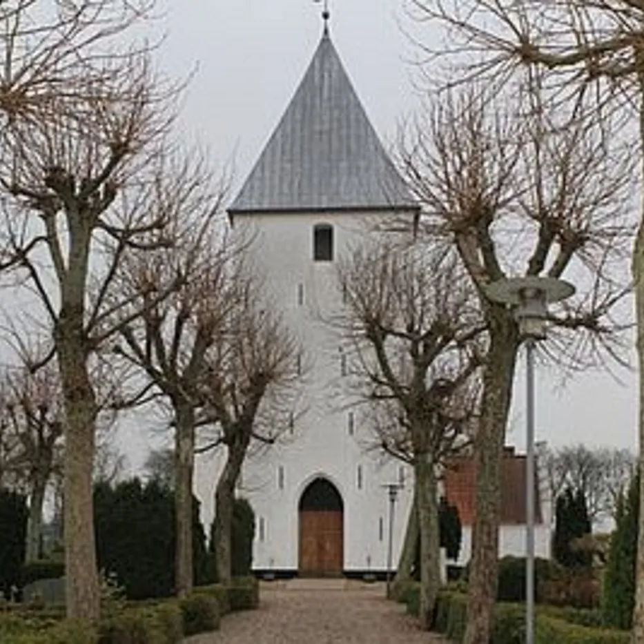 Hvid kirke med gråt spir set gennem nøgne træer, sti fører til dør.