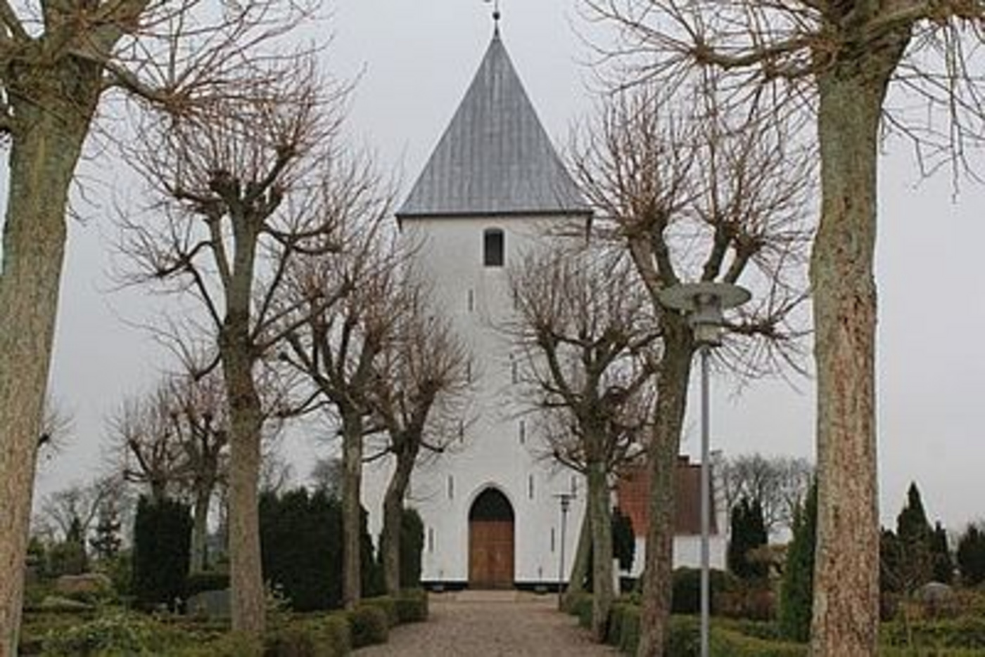 Hvid kirke med gråt spir set gennem nøgne træer, sti fører til dør.