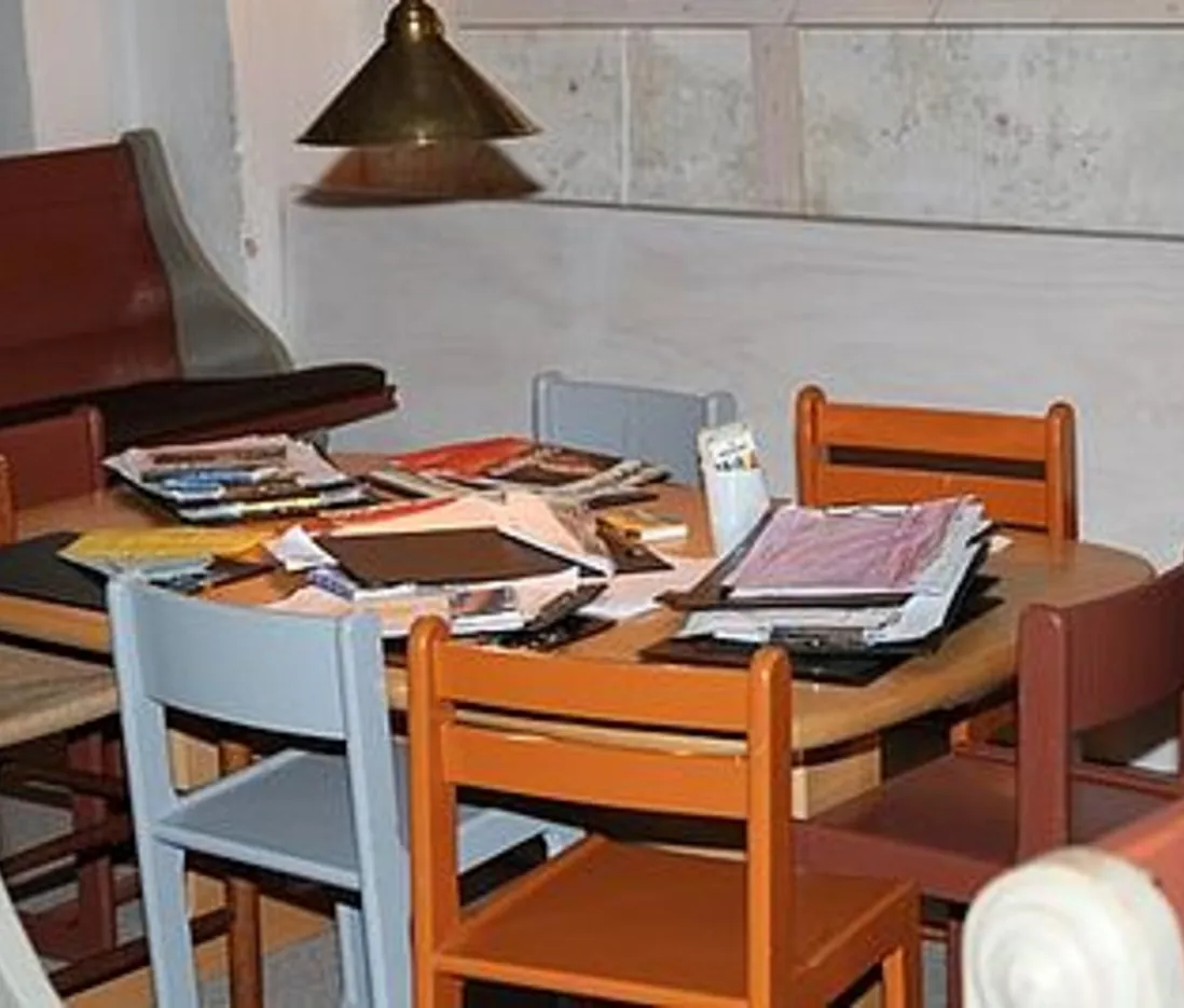 Rundt bord fyldt med papirer og bøger, omgivet af stole.