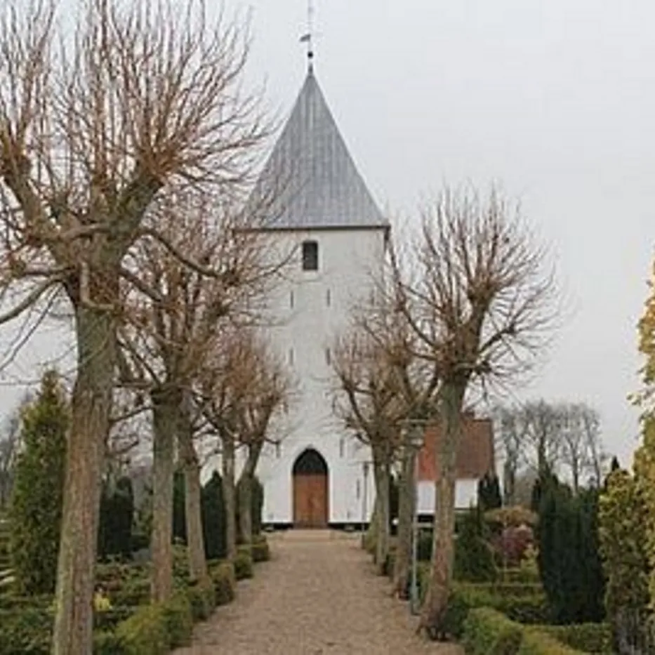 Hvid kirke med spir indrammet af nøgne træer, sti fører til indgangen.