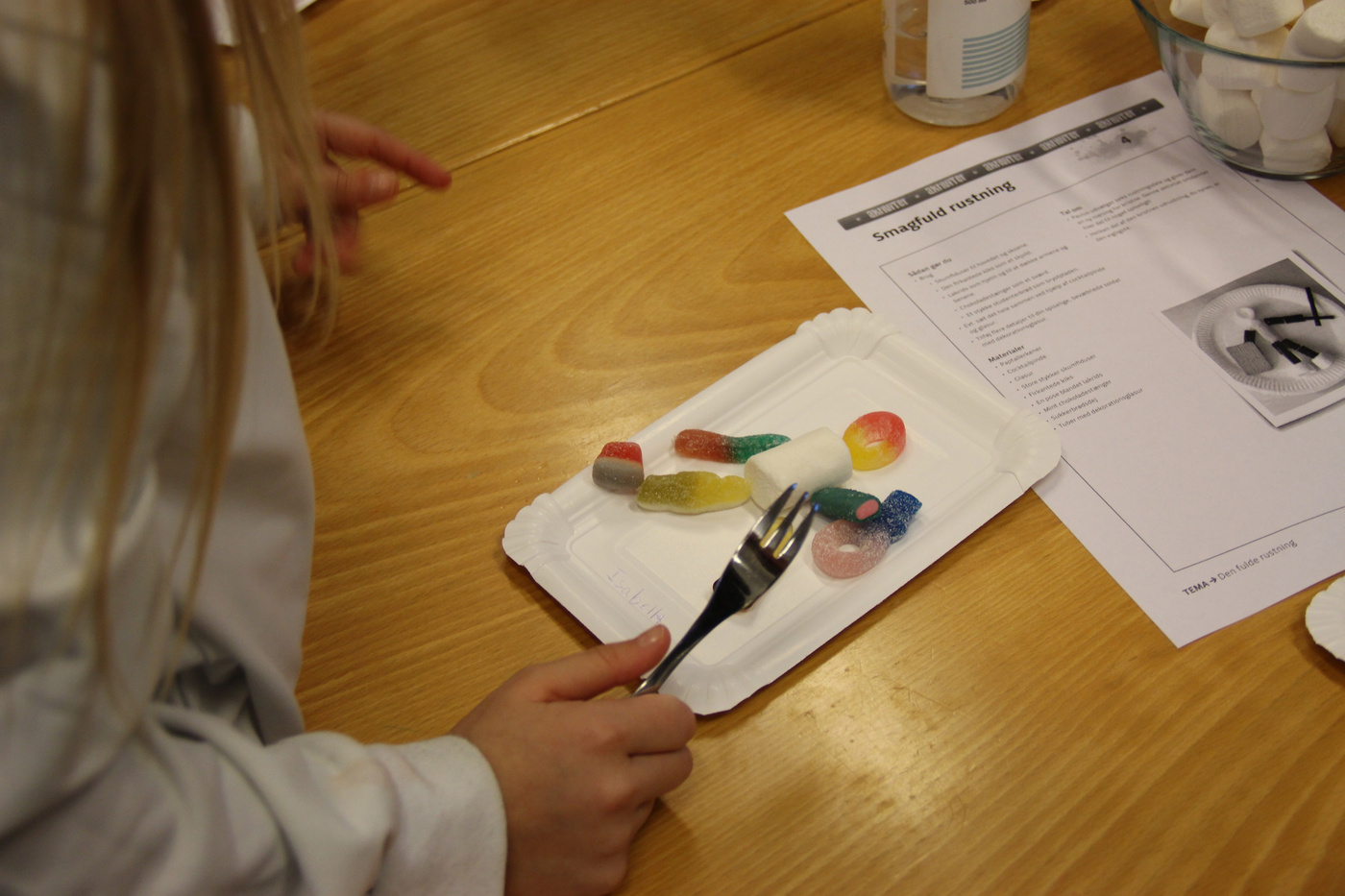  Barnehænder med gaffel, tallerken med slik og skumfiduser på bord.