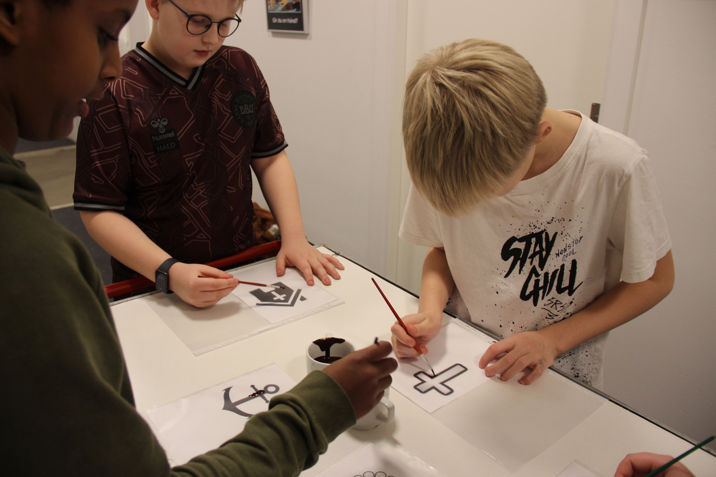 Børn maler designs, herunder et kors og anker, på papir.