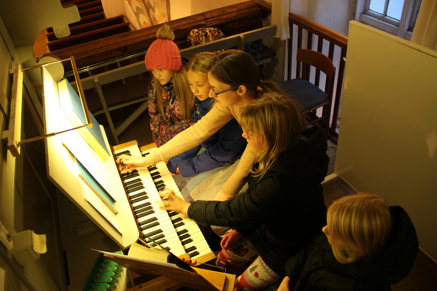 To piger spiller på kirkeorgel, overvåget af en voksen og et barn.