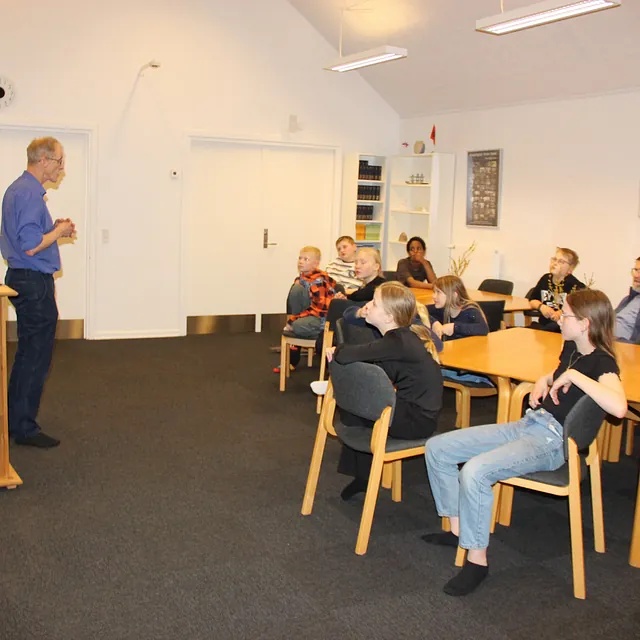 Mand står ved et talerstol og taler til et siddende publikum i alle aldre.