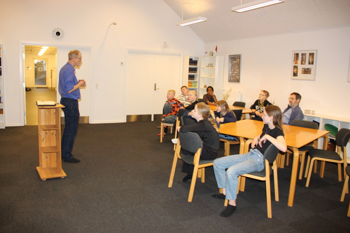 Mand står ved et talerstol og taler til et siddende publikum i alle aldre.