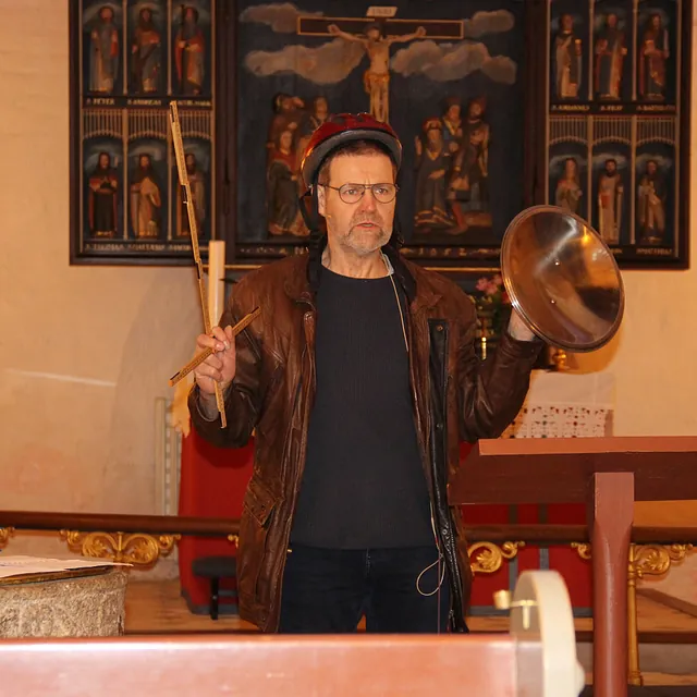 Mand med hjelm holder pinde og en bækken i en kirke.