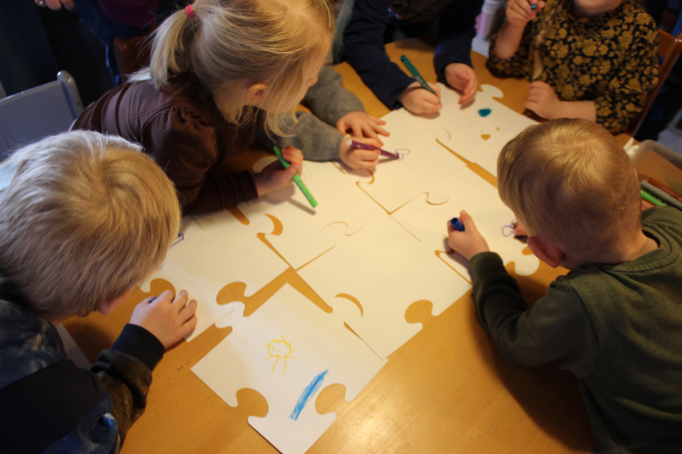 Børn farvelægger store puslespilsbrikker sammen ved et bord.