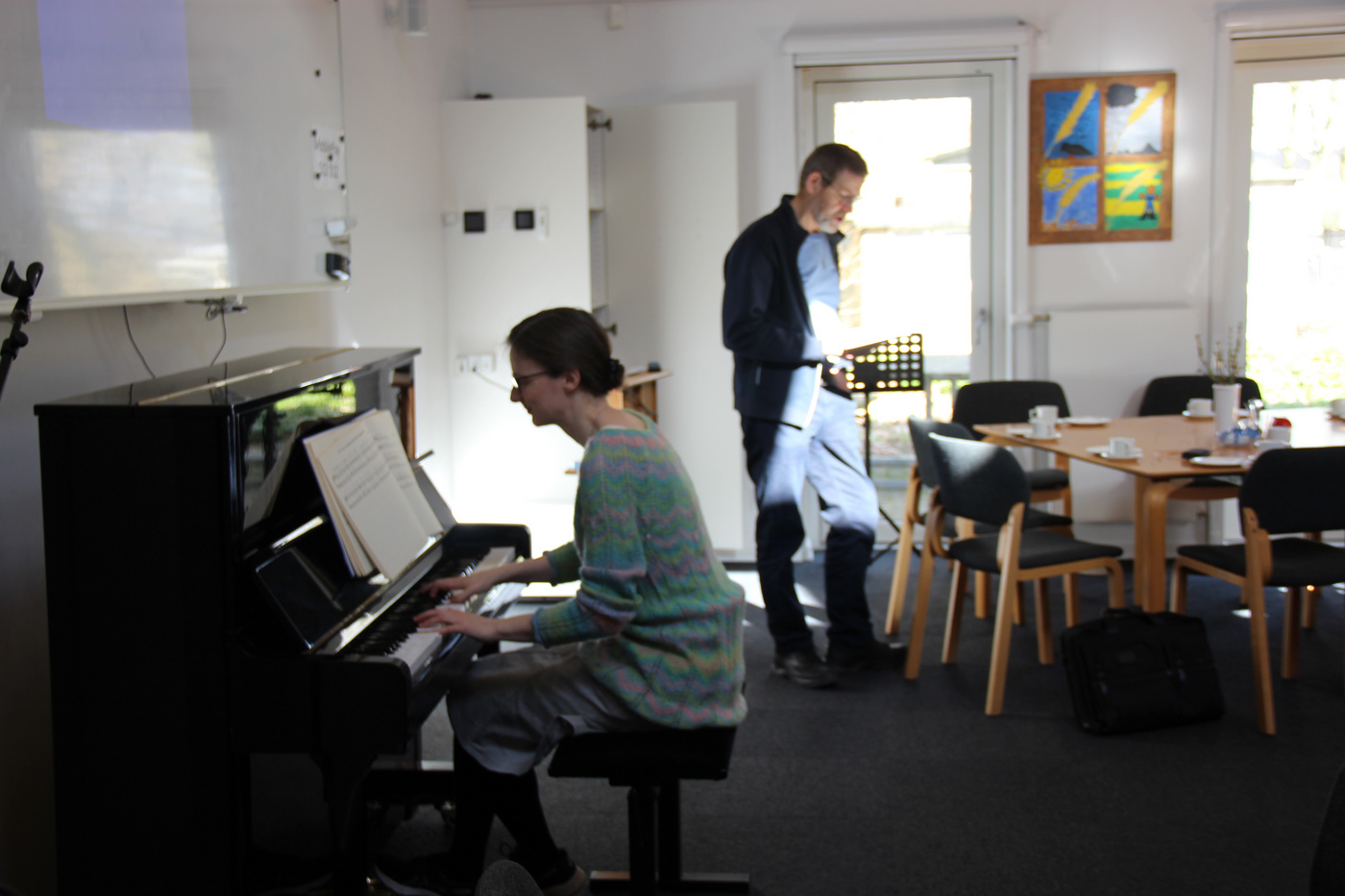Kvinde spiller klaver, mand står i nærheden i et lyst rum.