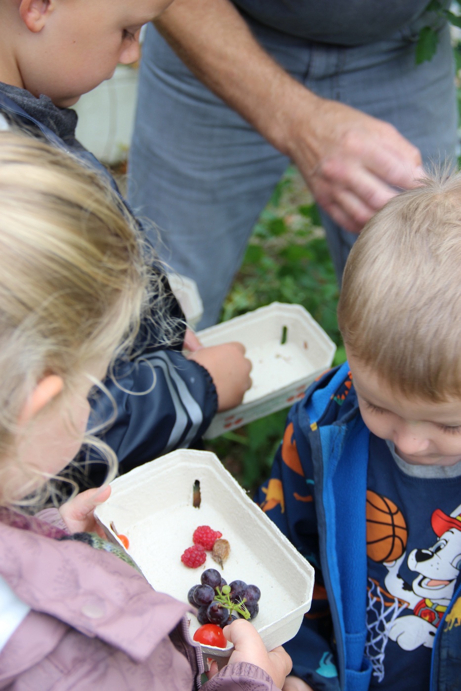 Børn holder bærbeholdere, voksne ben synlige i baggrunden.