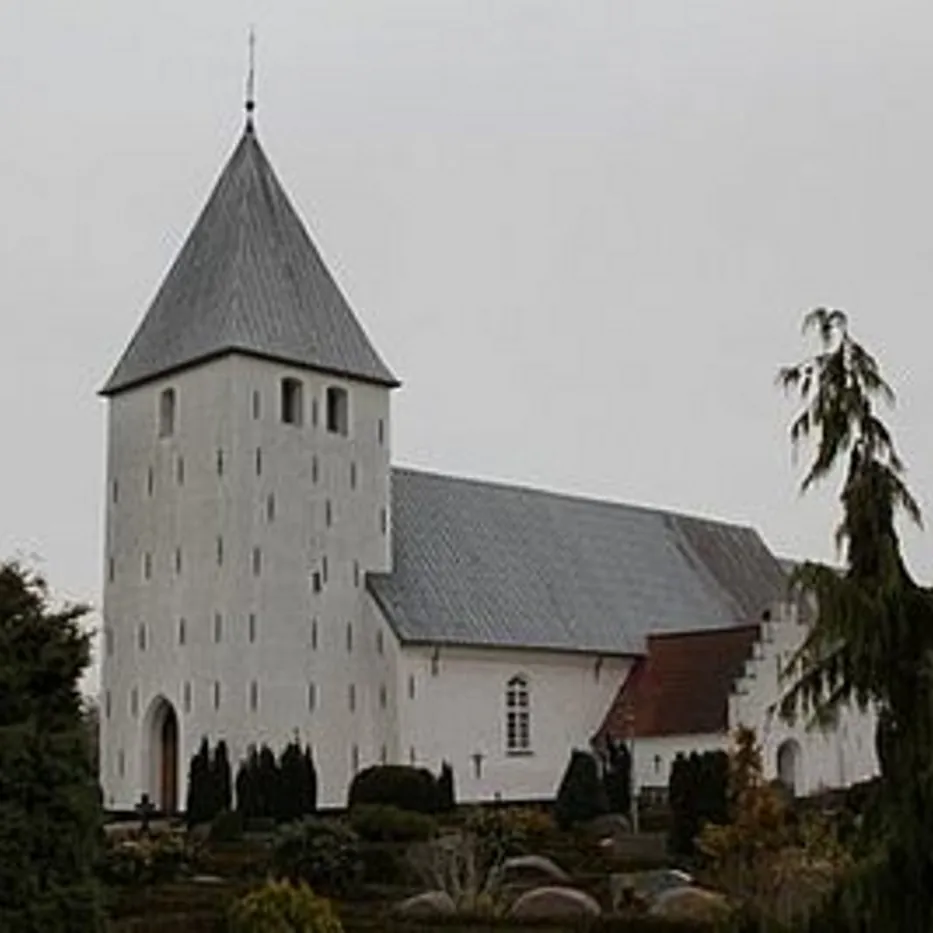 Hvid kirke med gråt spir og tag, omgivet af træer under en skyet himmel.