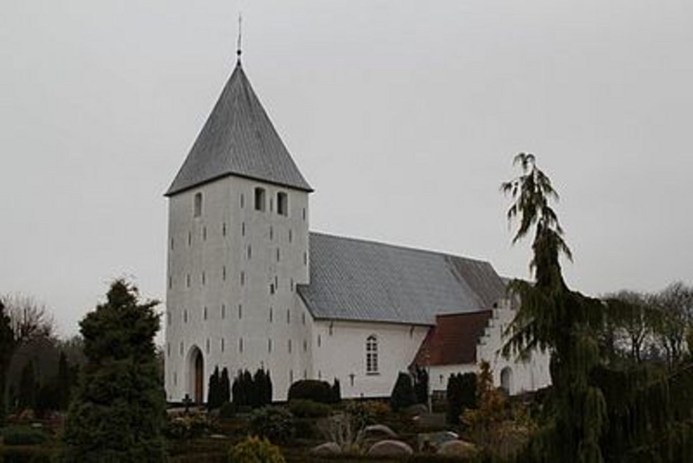 Hvid kirke med gråt spir og tag, omgivet af træer under en skyet himmel.