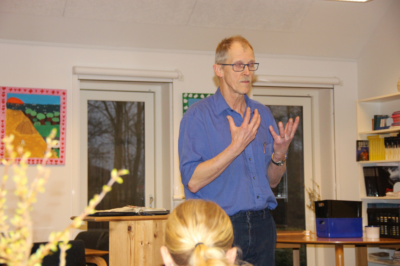 Mand med briller gestikulerer, mens han taler i et rum.