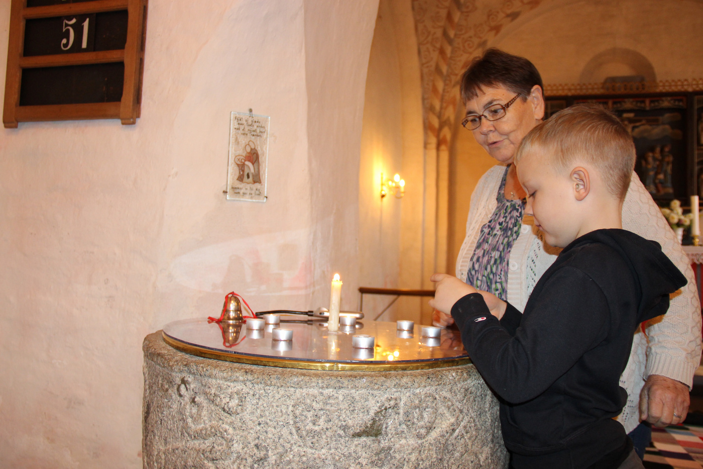 Dreng placerer et fyrfadslys på en stenfont, observeret af en kvinde.