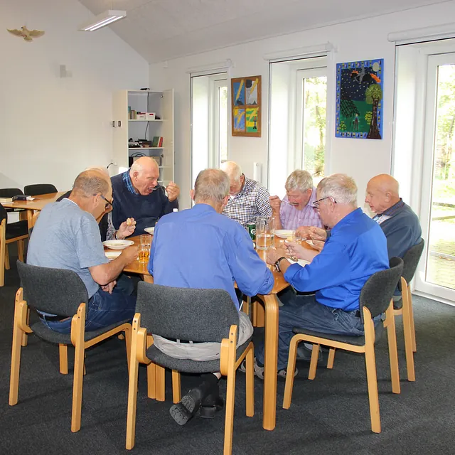 Ældre mænd samlet om et bord, spiser i et fællesrum.