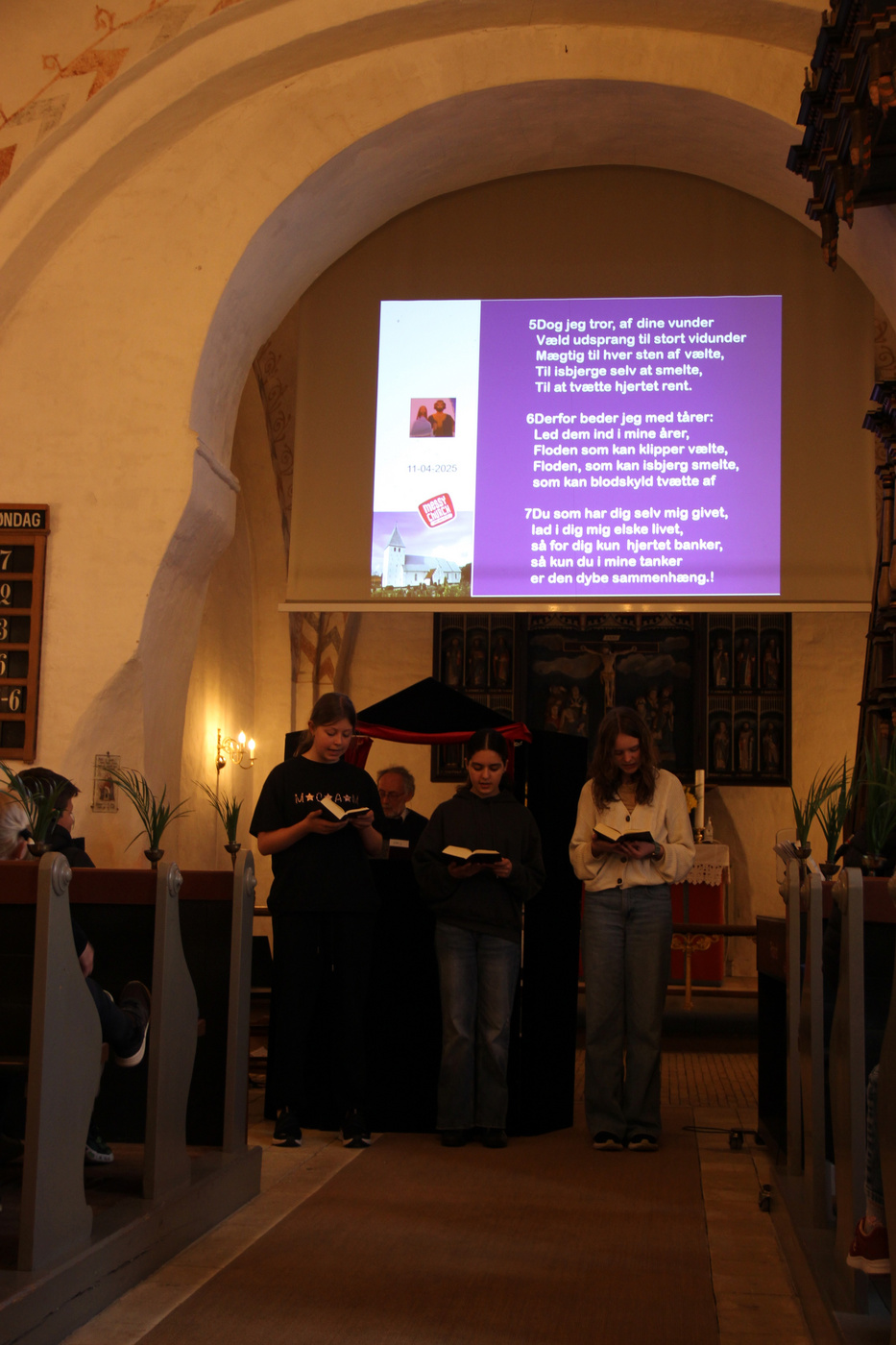 Tre unge læser fra bøger i en kirke med projicerede salmer.