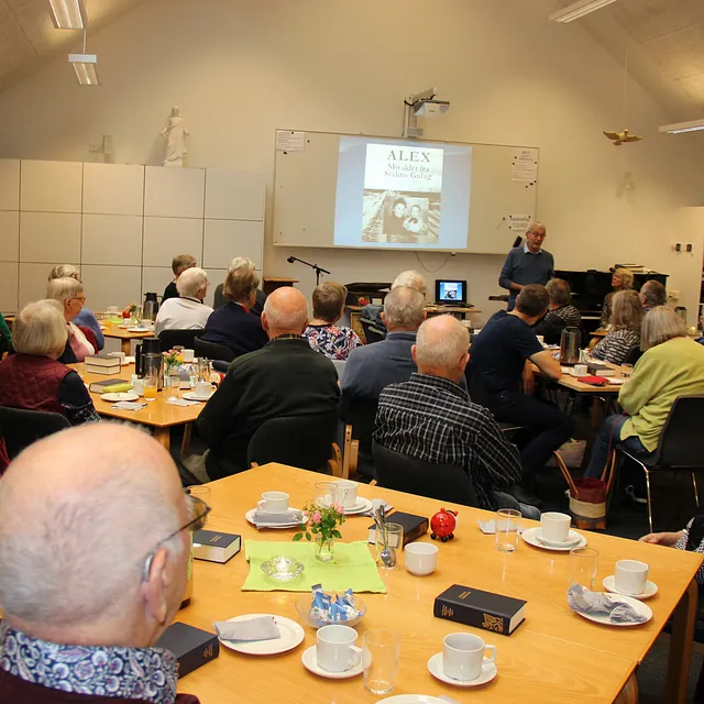 Stor gruppe voksne deltager i en præsentation i en kirkesal.