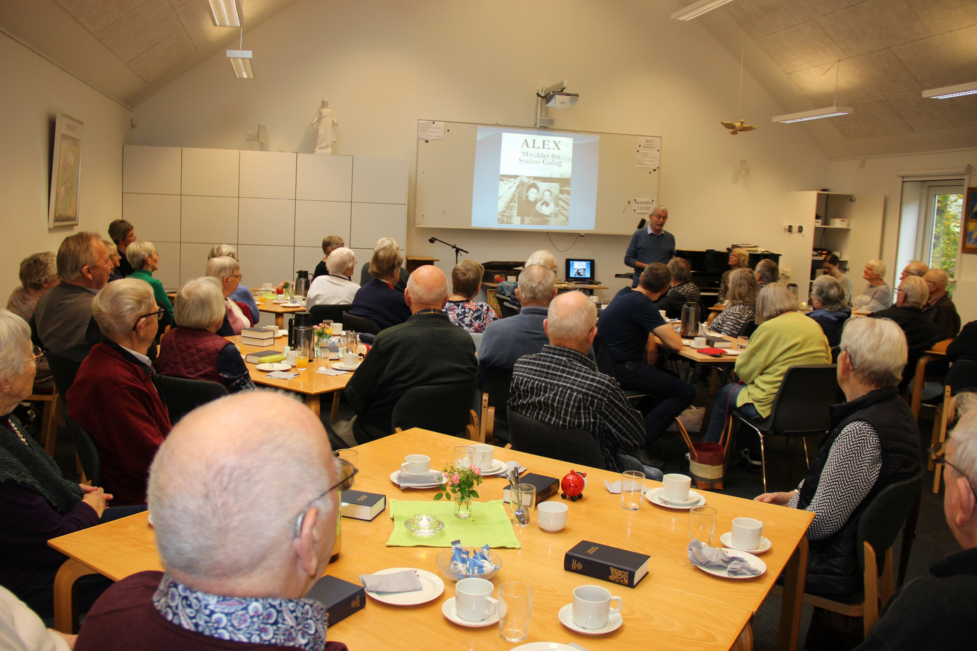 Stor gruppe voksne deltager i en præsentation i en kirkesal.