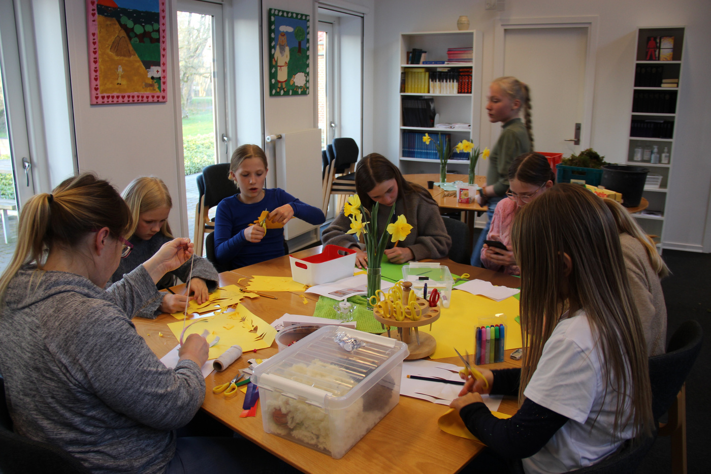 Børn og voksen laver håndarbejde med papir og saks ved et bord.