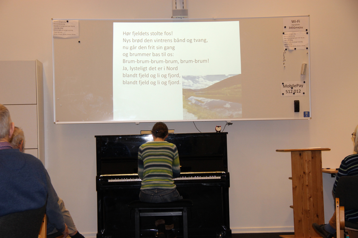 Person spiller klaver, vender mod skærm med danske tekster og landskab.