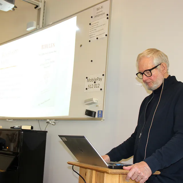 Mand med briller ved talerstol med bærbar computer, whiteboard bag ham.