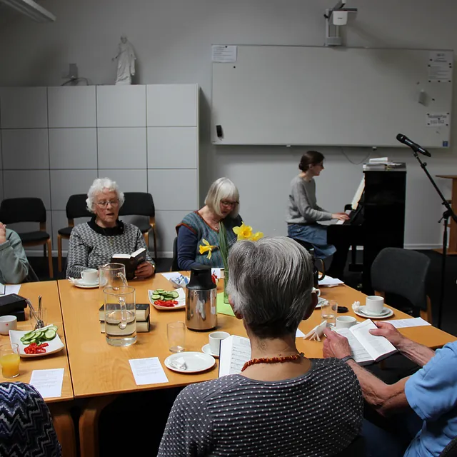 Ældre voksne læser og lytter omkring et bord, med en pianist i nærheden.