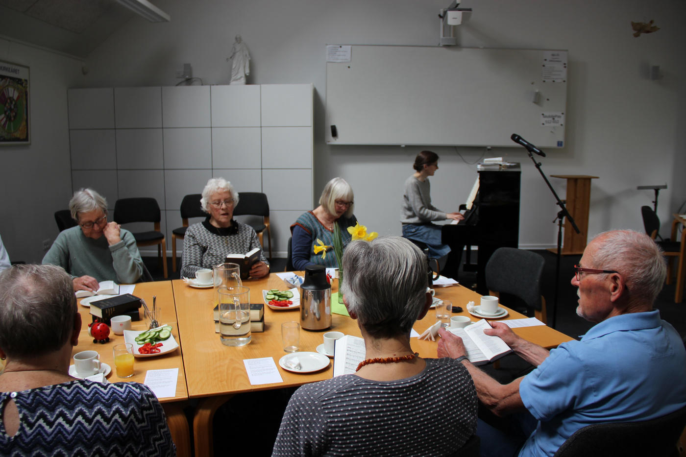 Ældre voksne læser og lytter omkring et bord, med en pianist i nærheden.