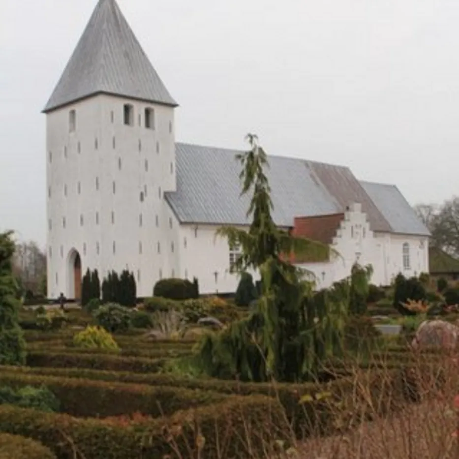 Hvid kirke med gråt tag og spir, omgivet af hække og træer.