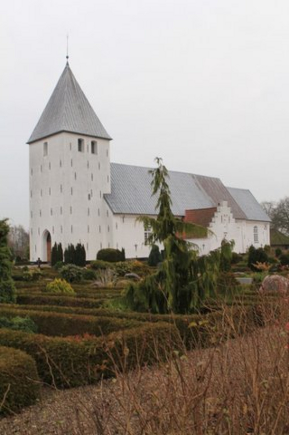 Hvid kirke med gråt tag og spir, omgivet af hække og træer.