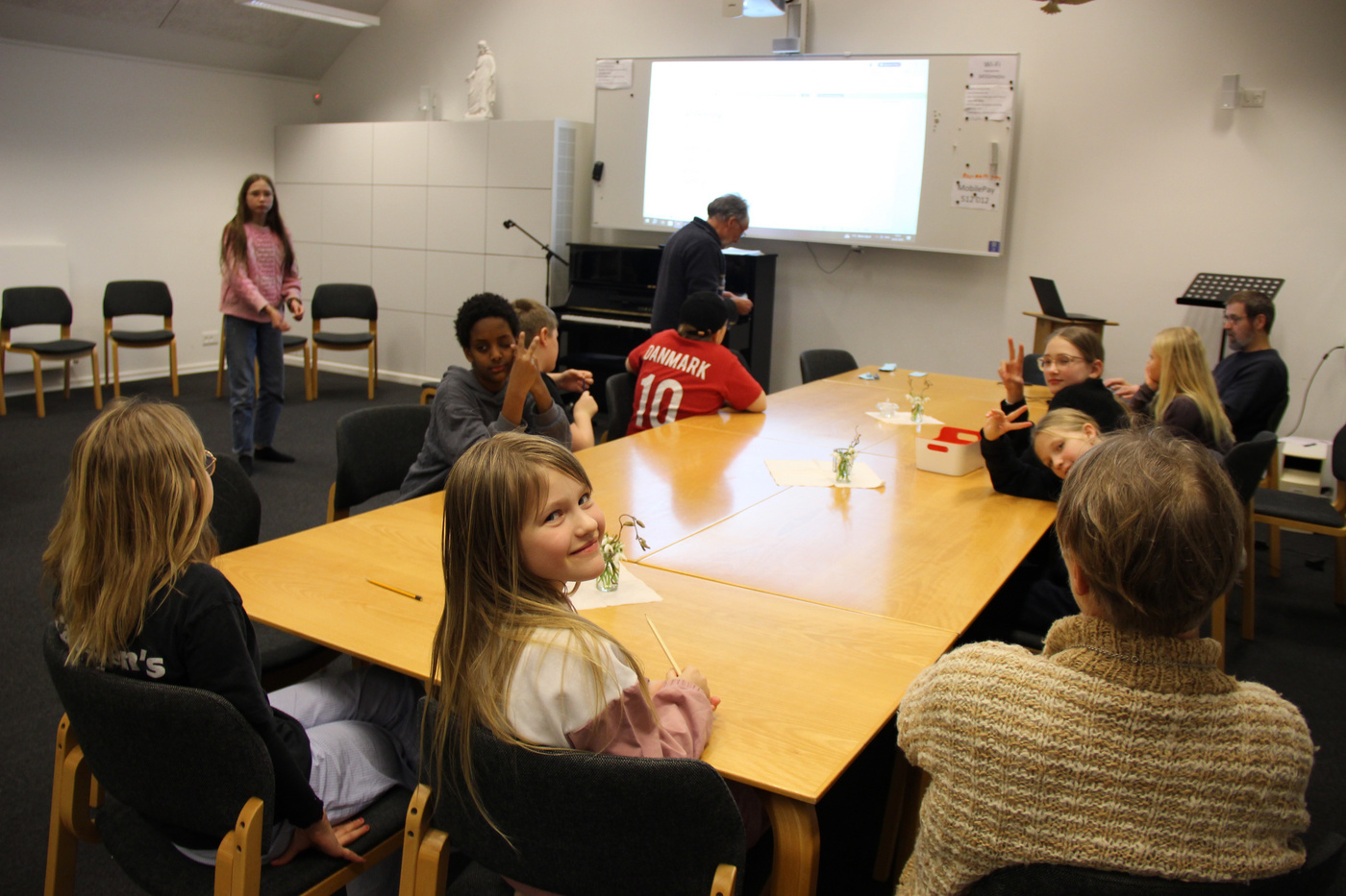 Børn og voksne samlet omkring borde i et fællesrum.