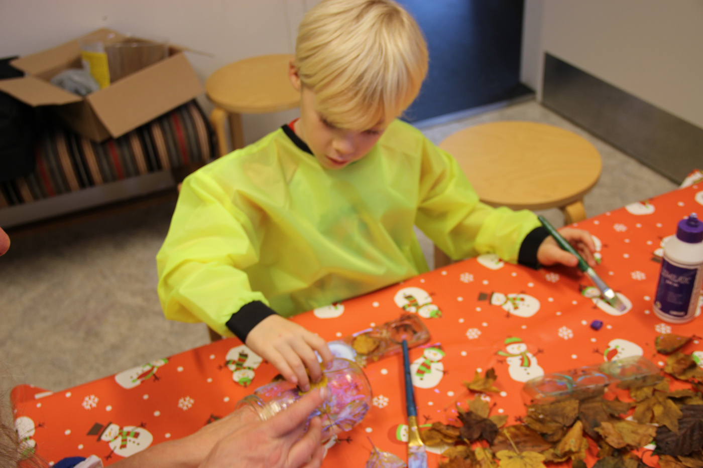 Barn i kittel maler en krukke ved et bord med efterårsblade.