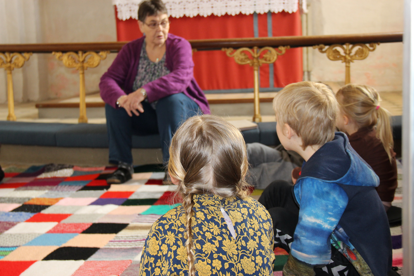 Kvinde taler til børn siddende på et farverigt kirkegulvtæppe.